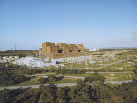 Aerial view of a colossal, earthy-toned industrial hydrogen energy complex juxtaposed against the stark white buildings and verdant trees under a serene sky, Sines, Setubal, Portugal.