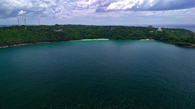 Wide aerial view of the Rumassala coastline with a calm bay, lush green hills, and cloudy sky, captured by a drone from high altitude.