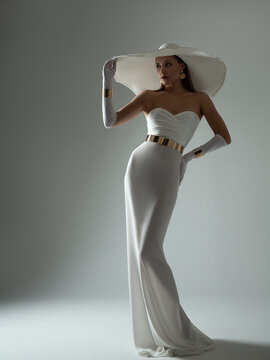Lady in White Dress and Wide Brim Hat. Wedding Fashion. Elegant Woman wearing evening Long Gown with Gloves Gray background