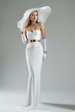 Lady in White Dress and Wide Brim Hat. Wedding Fashion. Elegant Woman wearing evening Long Gown with Gloves over Gray background