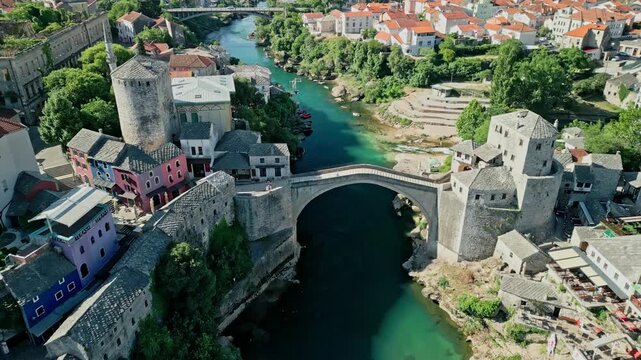 Angled forward dolly drone video showing Old Bridge Mostar with Old Bazaar, Kula Tara, Ćejvan-Ćehaja's Mosque, War Building, Halebija Tower, Neretva River, Lučki Most, and Mostar city along the river 