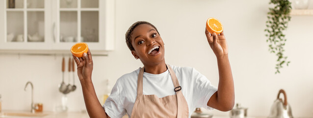 A woman stands in a kitchen holding two halves of an orange. She appears happy and engaged. The kitchen is bright with cabinets and utensils in the background. © Prostock-studio