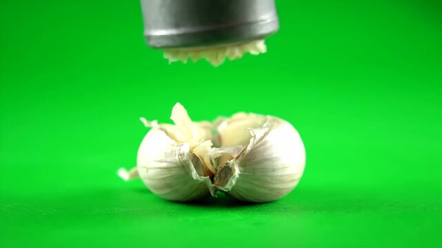 A metal garlic press crushes a fresh garlic bulb, extracting minced garlic onto a green screen surface. Ideal for cooking and food preparation visuals.