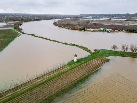 Aerial view of a lone, pale structure stands defiant amidst the floodwaters encroaching upon the verdant fields alongside the winding river, Portets, Nouvelle-Aquitaine, France.