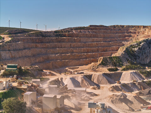 Aerial view of layered terraces carving into the earth, contrasting against the distant wind turbines, a stark reminder of industry's reach, Manilva, Andalusia, Spain.