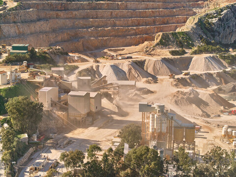 Aerial view of a vast, tiered quarry carved into the earth, heavy machinery dwarfed by the scale of the excavation, dust hanging in the air, Manilva, Andalusia, Spain.