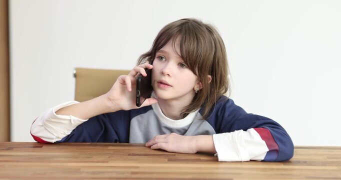 Girl talks on phone leaning on table against white wall at home after school break. Child scrolls screen watching actively after parents long call