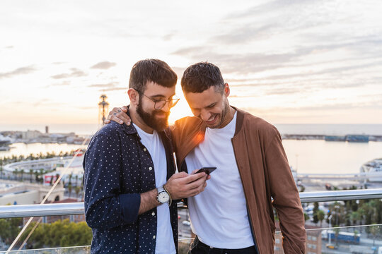 Gay couple using cell phone on lookout above the city with view to the port, Barcelona, Spain