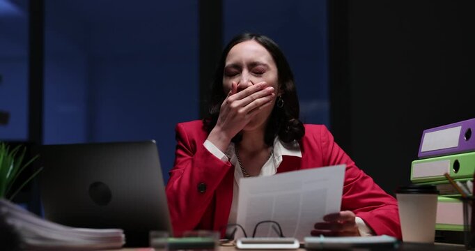 Businesswoman yawns covering mouth while reading report at night desk. Employee holds paper scanning laptop screen as overtime task continues