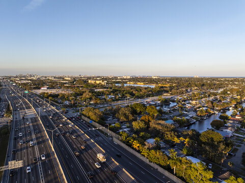 Aerial view of cars stream along the I-95 highway, cutting through a vibrant tapestry of green trees and sparkling waterways under a clear sky, Hollywood, Florida, United States.