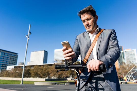 Mature businessman commuting in the city with his kick scooter, usine smartphone