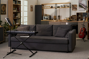 Modern living room featuring digital keyboard on stand with open sheet music and acoustic guitar leaning against kitchen counter, bookshelves and kitchen appliances visible in background © pressmaster