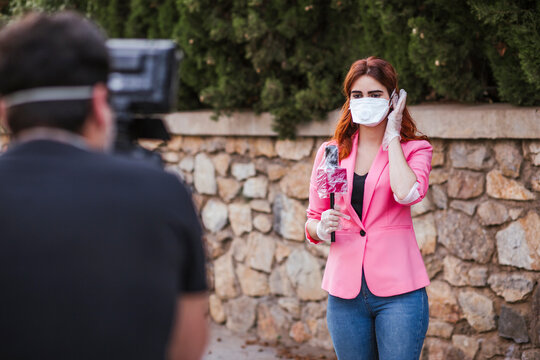 Camera operator filming reporter with microphone wearing mask while standing against wall