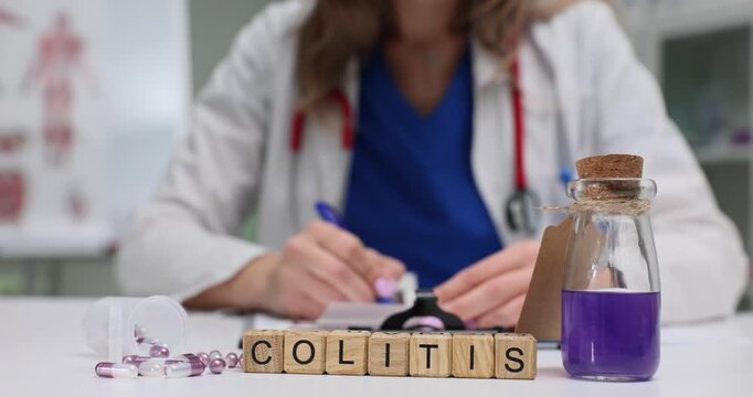 Wooden blocks spell word Colitis near medicine vial on clinic desk. Female doctor writes notes checking bottle label while forms treatment plan