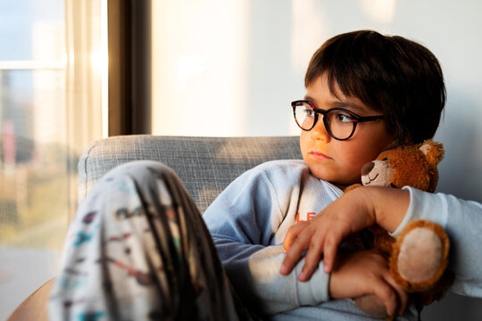 Portrait of serious boy with teddy bear sitting on armchair at home looking out of window