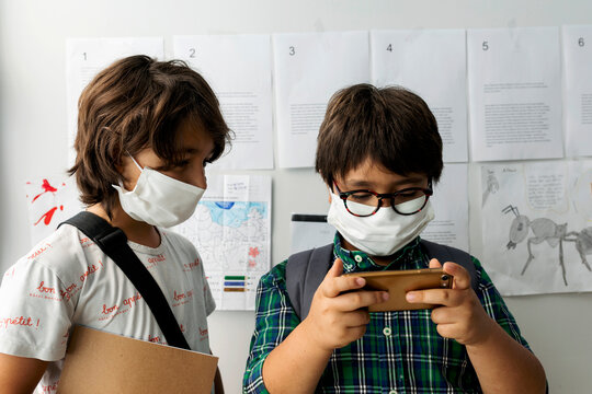 Boy wearing mask looking at friend using smart phone while standing against wall in school