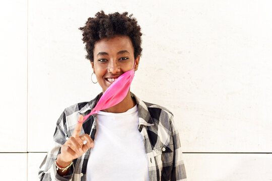 Smiling young woman removing pink mask against white wall
