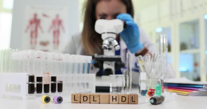 Wooden blocks spell abbreviation LDL HDL on laboratory desk. Young woman scientist uses microscope while checking blood sample tubes precisely