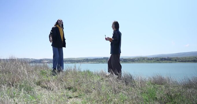 Female friends relax on river bank watching water and views under open sky. Women gesticulate and swirl laughing during weekend break outdoors