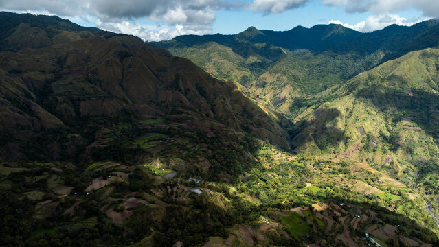 Aerial view of sunlight catching green valleys and ridged mountains under a partly cloudy sky, Igtuble, Tubungan, Philippines.