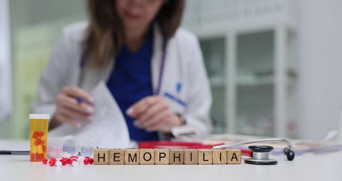 Wooden blocks spell word Hemophilia near scattered pills and stethoscope on clinic desk. Female doctor writes report checking bleeding disorder notes