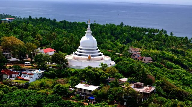Aerial view of the Japanese Peace Pagoda at Rumassala, a white stupa perched on a lush green hill above the coastline with trees, buildings and the ocean in the background.