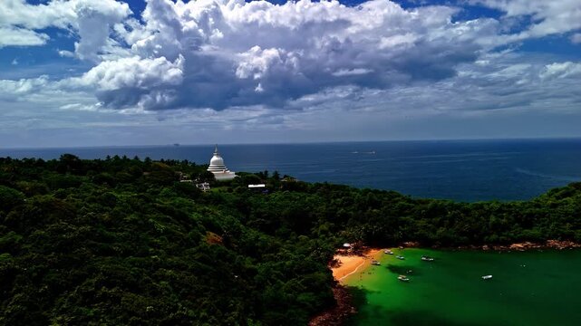 Aerial shot of the Japanese Peace Pagoda at Rumassala, perched on a lush green hill overlooking the ocean and beach with dramatic clouds.