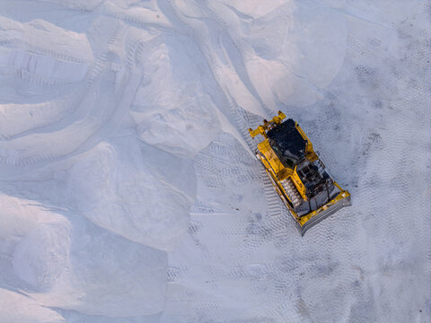 Aerial view of a yellow bulldozer carving paths through the stark white salt flats, creating a textured landscape of light and shadow, Aigues-Mortes, Occitanie, France.