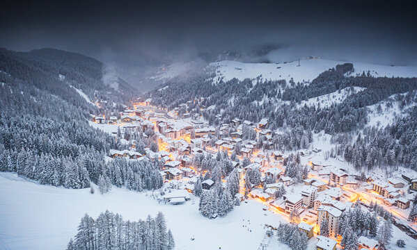 Aerial view of a snow-laden village of Madesimo nestled between dark forests, lights glowing warmly against the cold, crisp landscape, Madesimo Spluga Valley, Lombardy, Italy.
