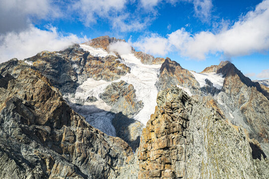 Aerial view of rugged, rocky slopes meeting glacial snow under a vibrant blue sky dotted with fluffy clouds, Monte Disgrazia, Valmalenco Sondrio, Lombardy, Italy.
