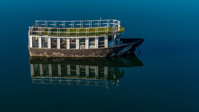 Aerial view of a weathered, two-story boat with a white-railed upper deck, sits still reflecting in the dark, glassy water., Knysna, Western Cape, South Africa.