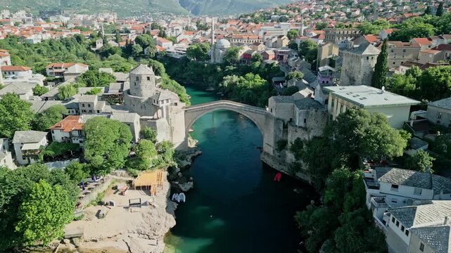 Going down drone video of Old Bridge Mostar in Old Town with Halebija Tower, Sultan Selimov Mesdžid, Koski Mehmed Pasha Mosque, Hajji Kurts Mosque, Neretva River, birds, and Beach below Stari Most on 