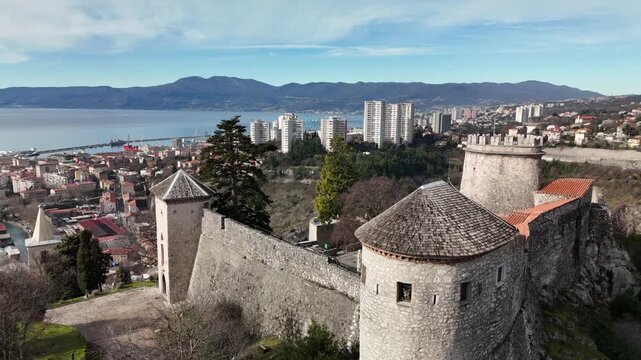 Drone lifts off beside Trsat Castle walls, rising to reveal the courtyard, hilltop fortress, Rijeka skyline, the Rječina valley, and the Adriatic Sea in the background.