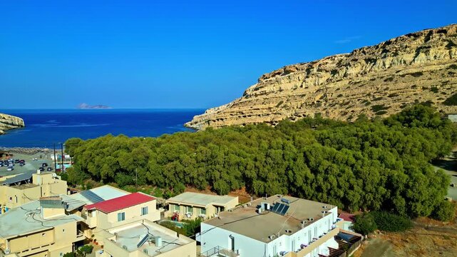 Flying Above Buildings And Trees With View Of Limestone Cliff Near Matala Beach In Crete, Greece. wide aerial shot