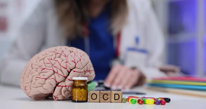 Miniature wooden cubes form abbreviation OCD near anatomical model of human brain. Young woman psychiatrist documents therapy plan at appointment
