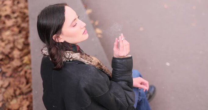 Young woman smokes cigarette enjoying calm atmosphere of autumn day. Female sits comfortably on park parapet and gazes into distance. Slow motion