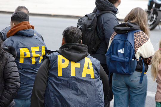 Argentine Federal Police officers (PFA vests) and civilians stand from behind in an urban setting.