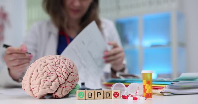 Abbreviation BPD formed from miniature wooden cubes near anatomical model of human brain on desk. Lady psychiatrist writes detailed recommendations