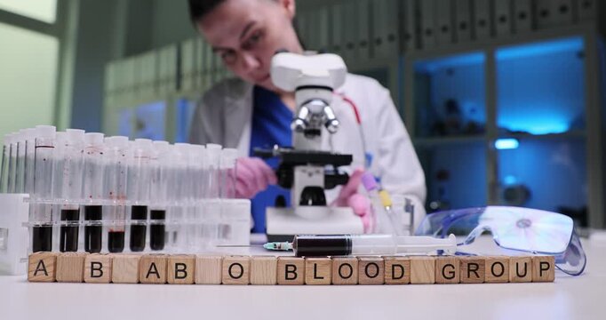Wooden cubes on laboratory table form letters A B AB O Blood Group by test tubes filled with blood samples. Lab worker examines blood under microscope