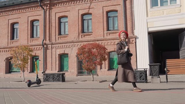 Young woman walking city street wearing red beret and brown coat, passing brick facade and wooden bench under autumn trees sequence shows student stride, tourist pause, street artist smile, candid
