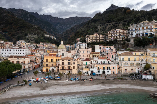 Aerial view of sun-kissed buildings cascade down to the sandy beach where colorful boats rest, framed by majestic mountains, Cetara, Campania, Italy.