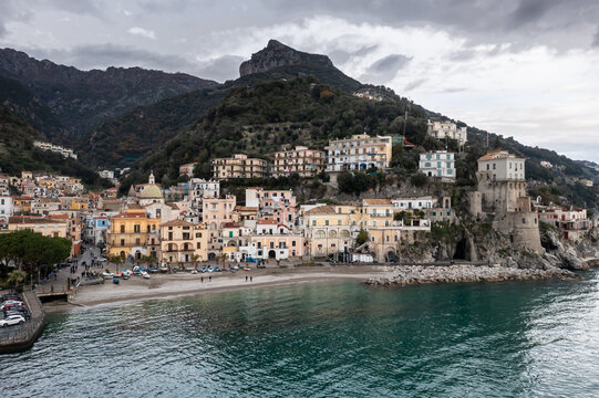 Aerial view of a charming coastal village nestled between the turquoise sea and rugged, green mountains, with a historic tower standing guard over the sandy beach, Cetara, Campania, Italy.