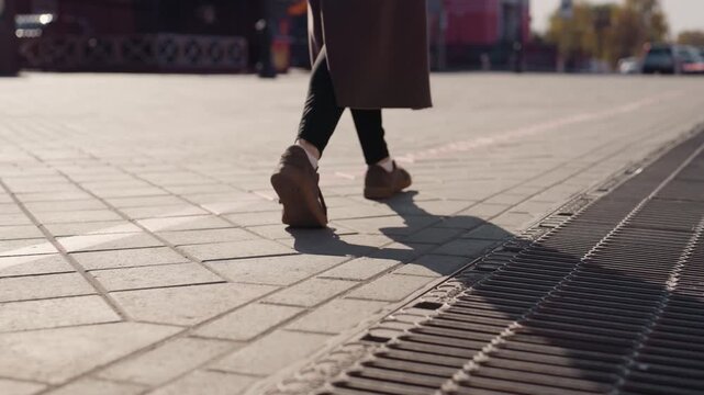 Ground level view walking on plaza person in long coat stepping over metal grate near tram lines sneakers scuffing patterned tiles electric scooter parked in background afternoon sunlight creating