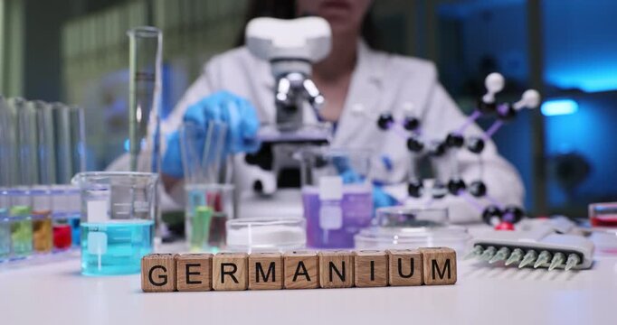 Wooden cubes on laboratory table form word Germanium showing element under study. Woman scientist adjusts microscope lens to observe details of sample