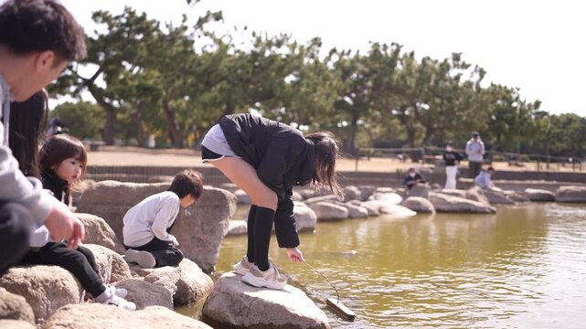 A group of people are sitting on rocks by a body of water. One woman is bending over and looking at something in the water