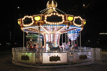Illuminated Paradise Carrousel shines brightly in a dark park at night, featuring horses and hot air balloon seats.