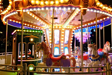 Brightly lit carousel with white horses, a green trolley, and a blue hot air balloon basket spins in an amusement park at night. © vannet