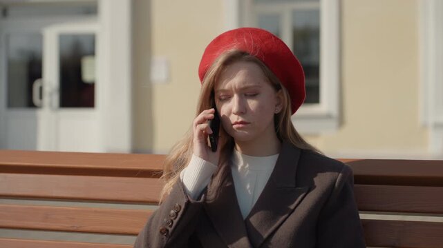 Young white woman bench checking phone, worried expression, red beret, wool coat, warm sunlight, pale city facade background, scrolling messages then answering call, quiet park bench scene,