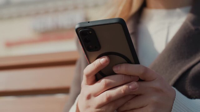 Hands holding smartphone on bench, woman in coat taps screen, sunlight glow, urban square background, closeup fingers scrolling messages, casual commuter, warm tones, shallow depth of field