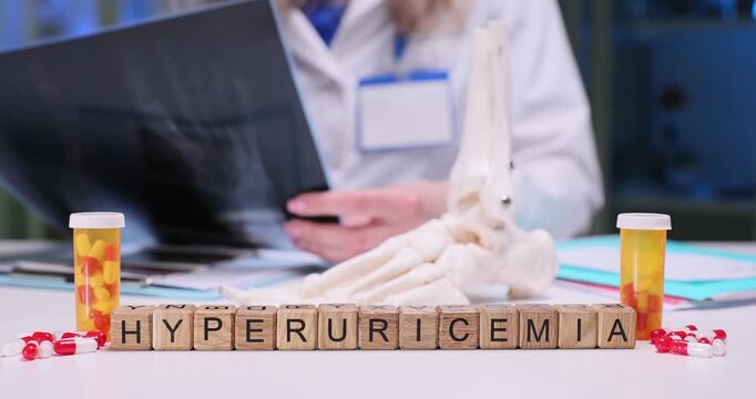 Wooden cubes on medical desk form word Hyperuricemia near anatomical model of foot and capsules. Woman doctor carefully studies X-ray image of feet